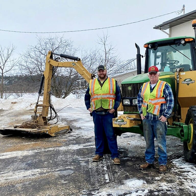 two guys standing in front of a tractor with a brush cutter